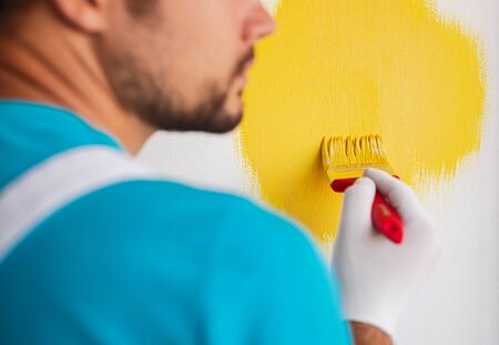 Back view of crop unrecognizable male worker with paintbrush painting wall in bright yellow color while decorating new apartmentの写真素材