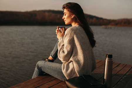 Calm woman with cup of hot drink sitting near lakeの写真素材