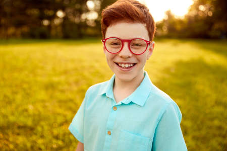 Excited redhead boy in trendy eyeglasses smiling at camera in natureの写真素材