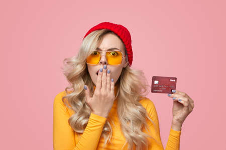 Shocked young woman covering mouth and demonstrating credit card in studio