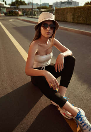 Trendy young lady sitting on asphalt road in sunlight