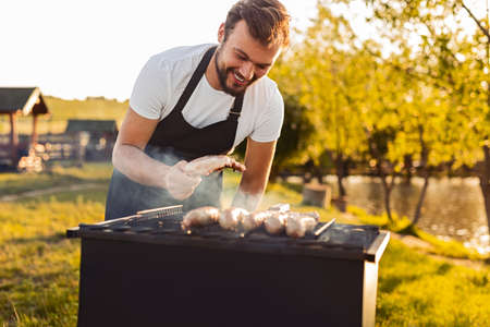 Cheerful man grilling sausages and meat in countryside