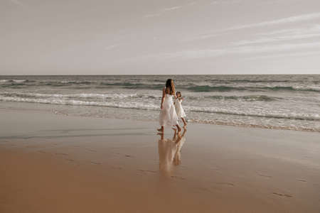 Mom and daughter in similar white dresses walking on sandy seashoreの写真素材