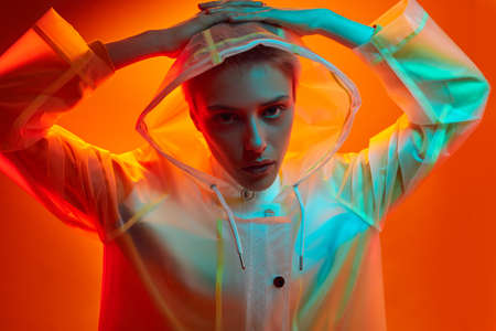 Serious young stylish woman in hooded raincoat touching head in studio
