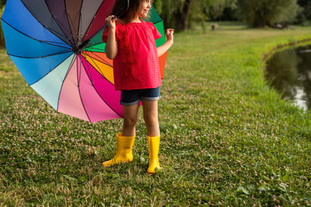 Adorable kid with rainbow umbrella standing on lawn in green parkの写真素材