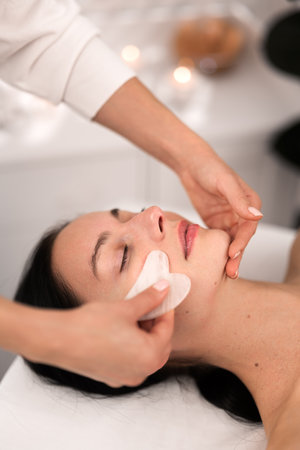 Crop beautician doing face massage with gua sha to female customerの写真素材