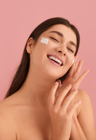 Excited young lady applying cream on face and smiling with closed eyesの写真素材
