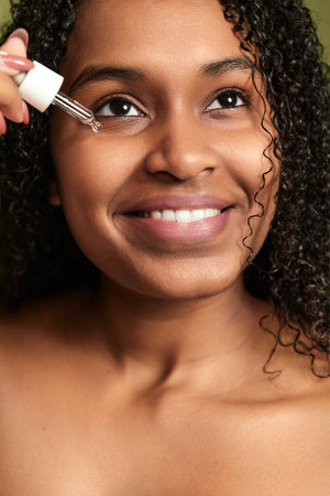 Smiling Hispanic female applying serum on face during skincare procedureの写真素材