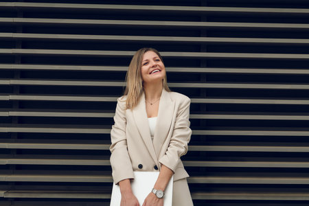 Joyful adult businesswoman with laptop smiling on city streetの写真素材