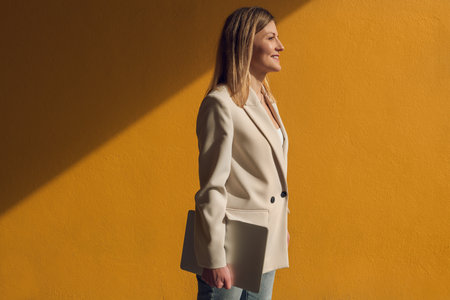Cheerful businesswoman standing against wall with laptopの写真素材