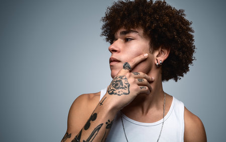 Serious young tattooed black man touching chin in gray studioの写真素材