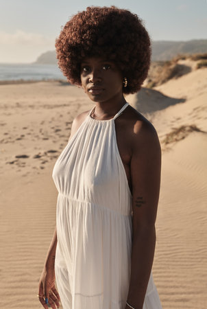 Young black woman in white dress standing on sandy beach in daylightの写真素材