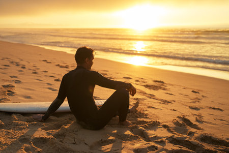 Surfer Sitting on Beach at Sunset with Surfboardの写真素材