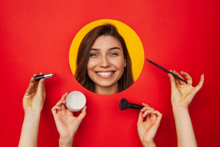 Smiling Woman Surrounded by Makeup Tools Against a Vibrant Red Backgroundの写真素材