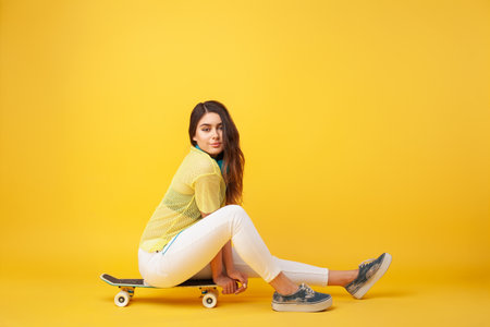 Casual Woman Sitting on Skateboard Against Vibrant Yellow Backgroundの写真素材