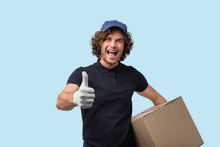 Smiling Delivery Worker With Thumbs Up and Package on Light Blue Backgroundの写真素材