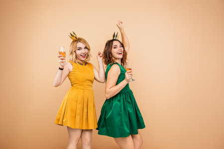 Two Women Celebrating in Elegant Dresses, Holding Glasses of Sparkling Drinksの写真素材