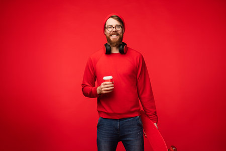 Cheerful Man in Red Outfit Holding Coffee Cup and Skateboard on Vibrant Backgroundの写真素材