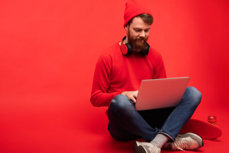 Young Man in Casual Outfit Working on Laptop in Vibrant Red Studioの写真素材