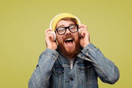 Excited Man Listening to Music on Headphones Wearing a Yellow Hat and Denim Jacketの写真素材