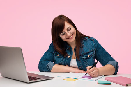 Smiling Woman Writing in Notebook by Laptop on Pink Backgroundの写真素材