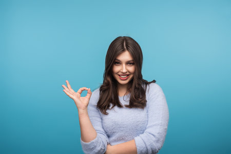 Smiling Woman Signaling Okay Gesture Against Blue Backgroundの写真素材