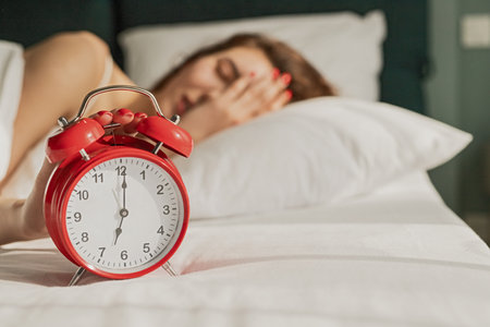 Woman Reaching for Red Alarm Clock While Lying in Bed in the Morningの写真素材