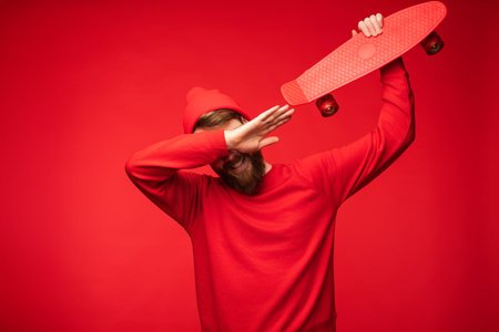 Young Man in Red Outfit Posing with Skateboard on Vibrant Red Backgroundの写真素材