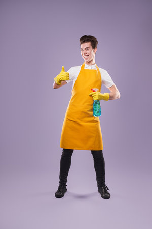 Happy Cleaner Wearing Yellow Apron Holding Spray Bottle and Gesturing Thumbs Upの写真素材