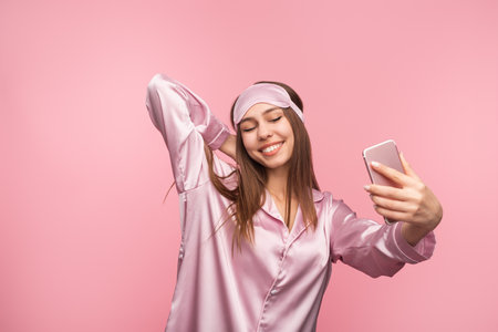 Cheerful Woman in Pink Pajamas Taking a Selfie Against a Pink Backgroundの写真素材