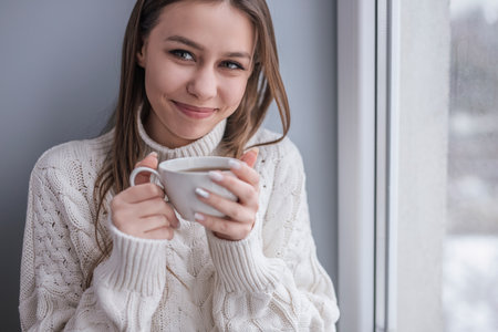 Smiling Woman in Warm Sweater Holding a Cup by the Windowの写真素材