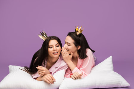 Two Friends in Pajamas Sharing a Secret on Bed with Crownsの写真素材