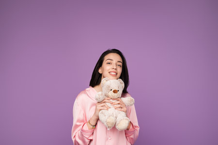 Young Woman Holding Teddy Bear Against Purple Background in Pajamasの写真素材
