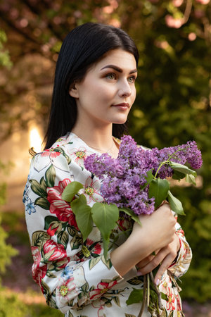 Woman Holding Lilac Flowers in Sunny Garden Wearing Floral Dressの写真素材