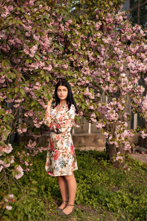 Elegant Woman in Floral Dress Posing Amid Blooming Cherry Blossoms Outdoorsの写真素材
