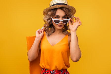 Stylish Woman Wearing Hat and Sunglasses Holding Shopping Bag on Yellow Backgroundの写真素材