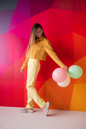 Stylish Woman Posing in Yellow Against a Vibrant Geometric Background Holding Balloonsの写真素材