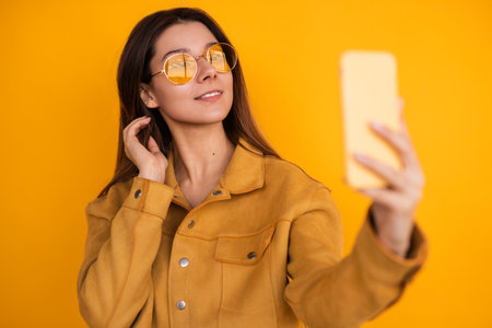 Stylish Woman Taking a Selfie Against a Vibrant Yellow Backgroundの写真素材
