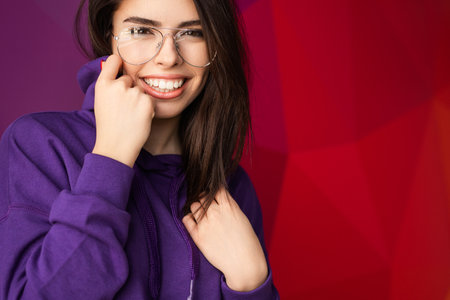 Smiling Woman in Glasses Wearing Purple Hoodie Against Vibrant Red Backgroundの写真素材