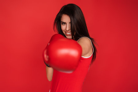 Confident Woman Wearing Boxing Gloves Against a Red Background Smiling Boldlyの写真素材