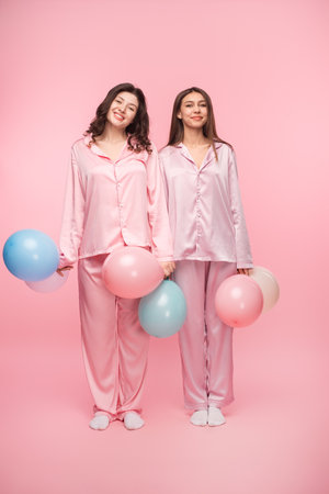 Two Friends in Pajamas Holding Colorful Balloons on Pink Backgroundの写真素材