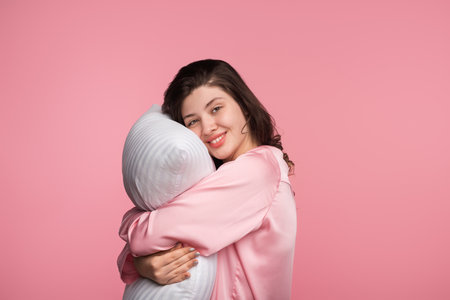 Woman in Pajamas Hugging Pillow on Pink Background Smiling Comfortablyの写真素材