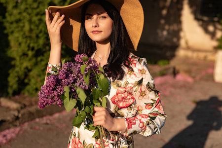 Elegant Woman Holding Purple Flowers in Sunlight Wearing Floral Dress and Hatの写真素材