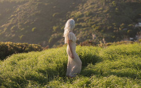 Woman in Flowing Dress Walking Through Sunny Green Meadow Landscapeの写真素材