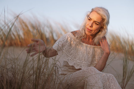 Elegant Woman in Lace Dress Sitting Amongst Beach Grasses at Sunsetの写真素材