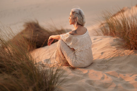 Peaceful Older Woman Enjoying a Serene Sunset on Sandy Dunesの写真素材