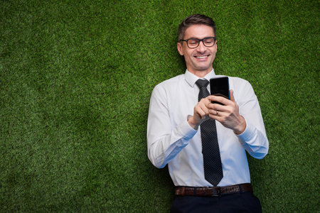 Smiling Professional Man Lying on Grass Using Smartphone in Formal Attireの写真素材