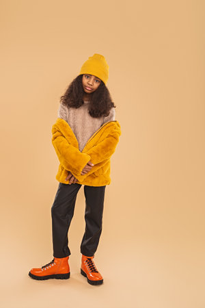 Stylish Young Girl in Yellow Beanie and Jacket Posing on Beige Backdropの写真素材