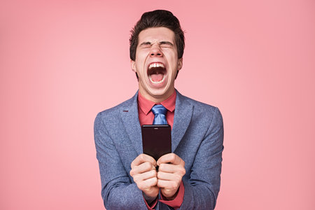 Frustrated Man Holding Smartphone Against Pink Background Expressing Strong Emotionsの写真素材