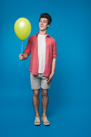 Cheerful Young Man Holding a Green Balloon Against a Bright Blue Backgroundの写真素材